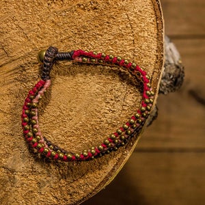 Puede incluir: Un brazalete trenzado rojo y marrón con cuentas doradas. El brazalete está sobre una superficie de madera.