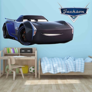 May include: A black and blue cartoon race car with a smiling face is on a light blue wall. The car is a wall decal with the name "Jackson" in a blue and silver shield above it. The car is in a child's bedroom with a wooden desk, chair, and bed.