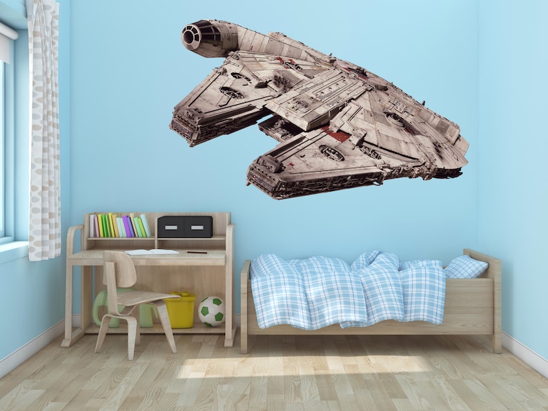 K&ouml;nnte beinhalten: Ein wei&szlig;er und grauer Raumschiff-Wandaufkleber mit einem roten Streifen, montiert an einer hellblauen Wand in einem Kinderzimmer. Der Aufkleber befindet sich &uuml;ber einem Holzbett mit einer blau-wei&szlig; karierten Bettdecke und einem Holzschreibtisch mit einem Stuhl und B&uuml;chern.