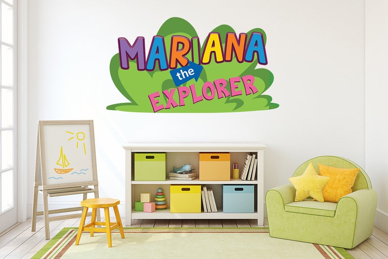 May include: A green, yellow, blue, pink, and purple wall decal with the text "Mariana the Explorer". The decal is on a white wall in a child's playroom. The room has a white bookshelf with colorful storage bins, a wooden easel with a drawing of a sailboat, and a green armchair with yellow pillows.