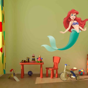 Op de afbeelding: Een groene en paarse zeemeermin-muursticker met rood haar en een paarse schelpbeha. De sticker zit op een lichtgroene muur in een kinderkamer met een rode tafel, een rode stoel en een speelgoed-driewieler.