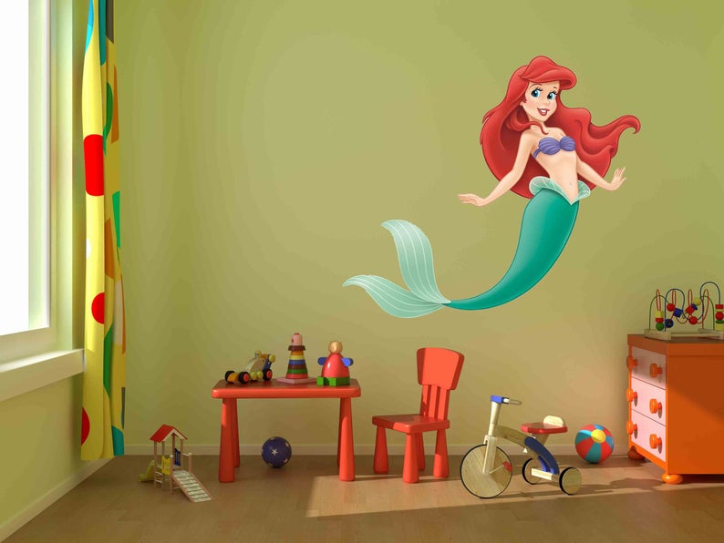 Op de afbeelding: Een groene en paarse zeemeermin-muursticker met rood haar en een paarse schelpbeha. De sticker zit op een lichtgroene muur in een kinderkamer met een rode tafel, een rode stoel en een speelgoed-driewieler.
