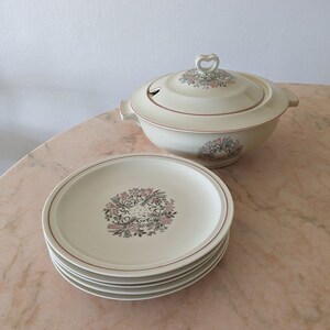 Peut inclure: Un ensemble de cinq assiettes en céramique blanche avec un motif floral dans des tons de rose, vert et marron. Les assiettes sont empilées les unes sur les autres. Un plat de service en céramique blanche avec un motif floral et une poignée en forme de cœur est en arrière-plan.