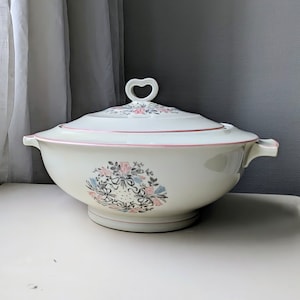 Peut inclure: Un bol de service en céramique blanche avec un motif floral rose et gris. Le bol a un couvercle avec une poignée en forme de cœur.