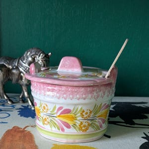 Puede incluir: Un azucarero de cerámica rosa y blanco con un diseño floral. El azucarero tiene una tapa y una pequeña cuchara de madera.