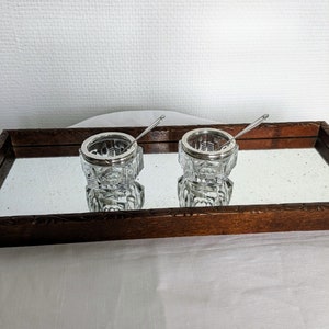 May include: Two clear glass salt cellars with silver lids and small spoons, sitting on a wooden tray with a mirrored surface.