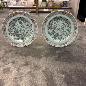 May include: Two matching decorative plates with a light teal background. Each plate features a floral and bird design in shades of blue, pink, and green. The plates have a decorative border with a repeating pattern. They are displayed on a gray countertop.