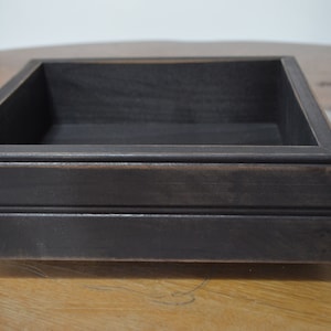 May include: A black wooden box with a square shape and a slightly distressed finish. The box has a simple design with a smooth surface and a raised edge around the top.