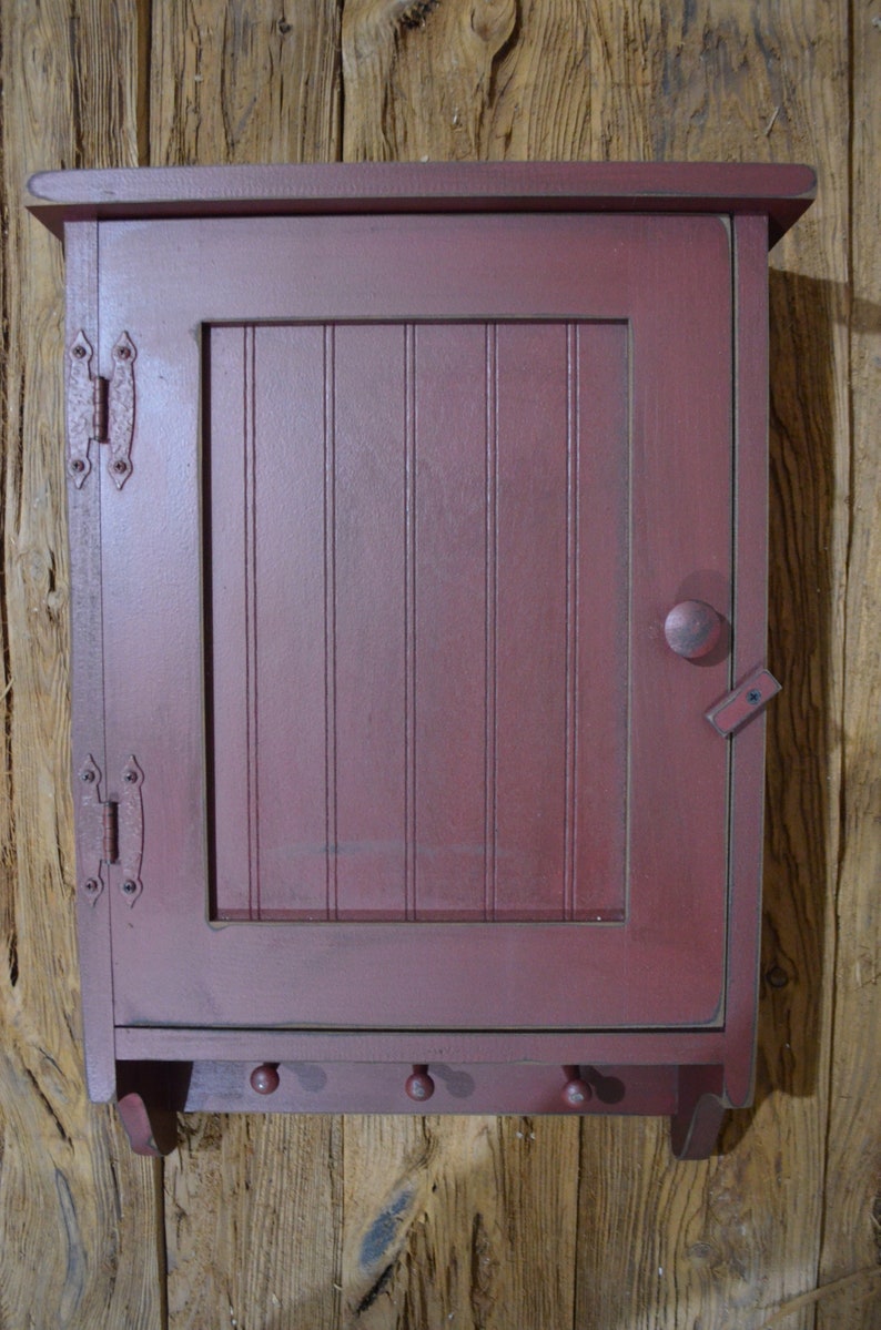 Puede incluir: Un peque&ntilde;o armario de madera rojo con una sola puerta y un estante. El armario tiene un acabado desgastado y presenta dos bisagras de metal y una perilla en la puerta. Hay tres ganchos en la parte inferior del armario.
