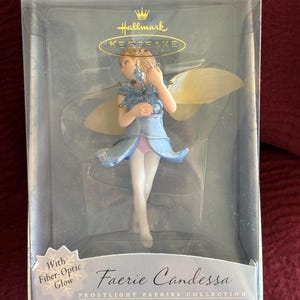 May include: A Hallmark Keepsake ornament featuring a fairy named Faerie Candessa. The fairy is dressed in a blue and pink dress with white tights and has translucent wings. The box reads "With Fiber-Optic Glow" and "Frostlight Faeries Collection."