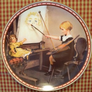 May include: A decorative plate featuring a painting of a young child painting a portrait of a doll. The plate has a red and white border and the artist's signature, "Norman Rockwell", is visible. The artwork depicts a child in a black dress and white collar.