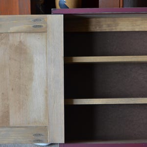 Puede incluir: Un peque&ntilde;o armario de madera con un exterior pintado de rojo y una puerta de madera natural. El armario tiene tres estantes en el interior.