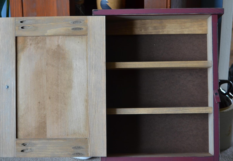 Puede incluir: Un peque&ntilde;o armario de madera con un exterior pintado de rojo y una puerta de madera natural. El armario tiene tres estantes en el interior.