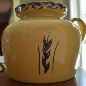 May include: A yellow ceramic pitcher with a blue and white floral design. The pitcher has a handle and a spout. It has a wheat design on the front.