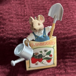 May include: A decorative ornament featuring a mouse wearing blue overalls, holding a silver watering can and a shovel. The mouse is positioned inside a rectangular packet labeled STRAWBERRIES, which displays a graphic of red strawberries.
