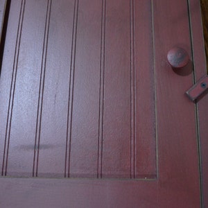 Puede incluir: Una puerta de armario de madera roja con un pomo simple y un pestillo de metal. La puerta tiene ranuras verticales y un acabado desgastado.