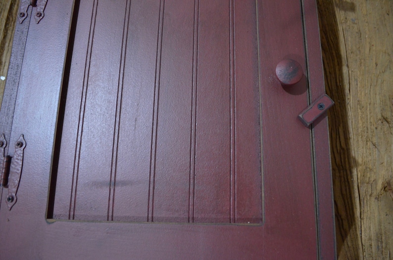 Puede incluir: Una puerta de armario de madera roja con un pomo simple y un pestillo de metal. La puerta tiene ranuras verticales y un acabado desgastado.