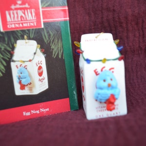 May include: A white ceramic egg nog carton Christmas ornament with a blue bird peeking out of the top. The carton has a red and white label that says "Egg Nog" and has a string of colorful lights around the top. The ornament is in its original box, which says "Keepsake Ornament" and "Egg Nog Nest".