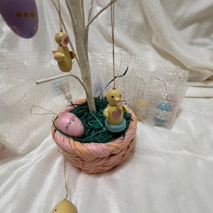 May include: A decorative Easter tree with hanging ornaments. The tree features a woven basket base filled with green faux grass and small wooden ducklings. Painted Easter eggs in various colors are also hanging from the branches.