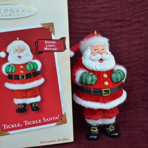 May include: A red and white Santa Claus ornament with a white beard and green mittens. The ornament has a light-up feature and plays a sound. The box reads "Tickle, Tickle Santa!" and "Sound, Light, Motion!"