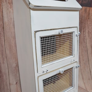 May include: A white, distressed-finish cabinet with a sloped top and two wire-mesh fronted compartments. The cabinet has a handle on top and small knobs on the doors. The cabinet is made of wood and has a rustic, farmhouse style.