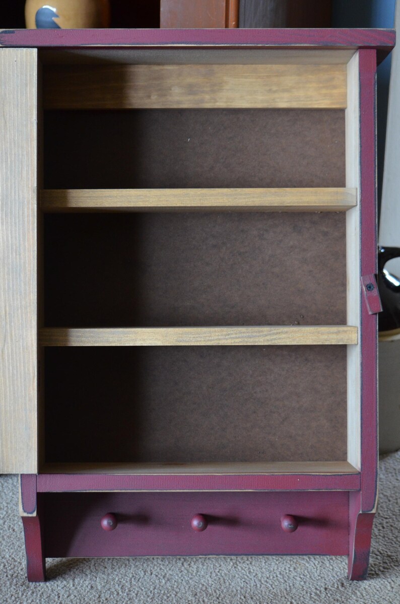 Puede incluir: Una peque&ntilde;a estanter&iacute;a de pared de madera con un acabado rojo burdeos. La estanter&iacute;a tiene tres estantes y dos ganchos en la parte inferior. La estanter&iacute;a est&aacute; vac&iacute;a.