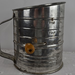 May include: A vintage silver-colored metal flour sifter with a wooden handle. The sifter has markings for two, three, four, and five cups.