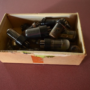 May include: A cardboard box filled with vintage vacuum tubes. The tubes are made of glass and metal and have a variety of shapes and sizes.