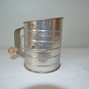 May include: A silver metal flour sifter with a wooden handle. The sifter has markings for one, two, and three cups.