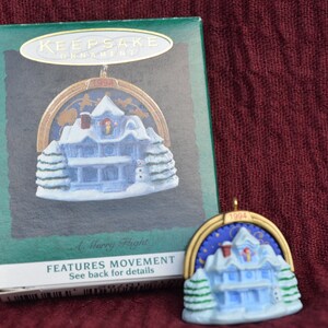 May include: A blue and white Christmas ornament featuring a house with a snowy roof and a moving light inside. The ornament is in its original box, which has the text "Keepsake Ornament" and "Features Movement" on it. The ornament is from 1994.