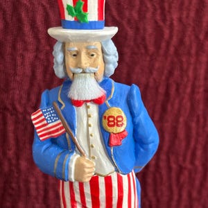 May include: A figurine of Uncle Sam, dressed in a blue coat, red and white striped pants, and a tall hat. He holds a small American flag and has a white beard and mustache. The figurine has a gold '88' pin.