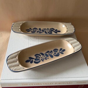 Peut inclure: Deux plats en céramique de forme ovale allongée avec une base de couleur beige. Chaque plat présente un motif floral bleu foncé au centre et des détails texturés en forme de vannerie aux extrémités arrondies, avec un fin bord bleu.