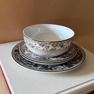 May include: A white ceramic bowl and two plates with a black and beige floral design. The bowl and plates are stacked. The plates and bowl have a black border. The set is on a white surface.