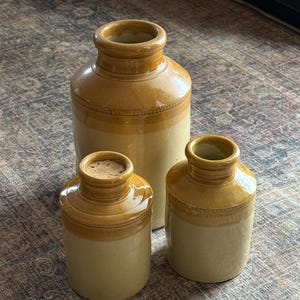 Peut inclure: Trois pots cylindriques en grès avec une finition bicolore, présentant une glaçure brillante brun miel sur la partie supérieure et une base mate de couleur crème. Les pots ont une ouverture à bord épais et un petit bouchon en liège.
