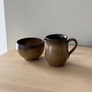 May include: A brown ceramic jug and bowl set. The jug has a handle and spout, and the bowl is round. Both items have a gradient colour, transitioning from a lighter brown to a darker brown.