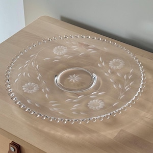 May include: A clear glass serving platter with an etched floral and leaf design. The platter has a raised centre and a beaded edge. The floral design includes flowers and leaves. The platter is on a wooden surface.