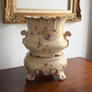 May include: A cream-coloured ceramic jardiniere with a matching pedestal base, featuring delicate hand-painted floral motifs in pink, purple, and green. The vessel has a scalloped rim, ornate side handles, and a crack visible on the upper body.
