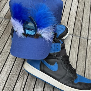 May include: A blue baseball cap adorned with blue feathers and a decorative stone, paired with a black and blue high-top sneaker. The sneaker features a blue swoosh and a white sole, creating a stylish ensemble.