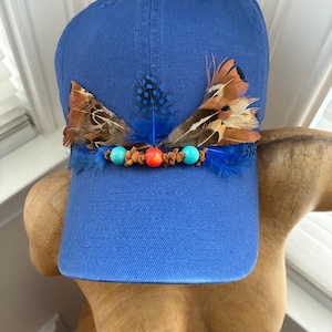 May include: A blue baseball cap adorned with feathers and beads. The cap features brown and orange feathers, blue feathers, and turquoise and orange beads. The hat is displayed on a wooden form.