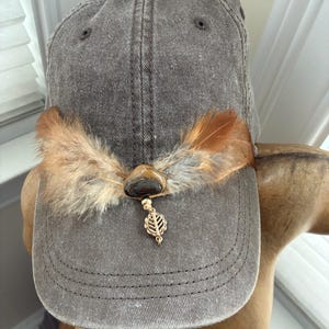 May include: A gray baseball cap with a decorative feather and stone embellishment. The cap features a brown stone pendant with a gold-colored leaf charm. The feathers are brown and tan.