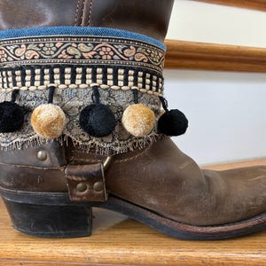 May include: Brown leather boot with decorative trim. The trim includes a blue denim band, patterned fabric, black and cream-colored pom-poms, and lace. The boot has a square toe and a low heel.