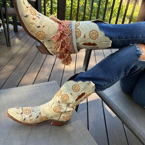 Può includere: Stivali da cowboy color crema con ricami floreali nei toni dell'arancione, giallo e blu. Gli stivali hanno la punta a punta e un polsino decorativo alla caviglia con nappe. Gli stivali sono indossati con jeans blu.