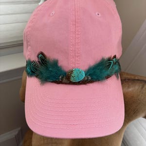 May include: A pink baseball cap with a turquoise stone and teal feathers. The cap has a curved brim and a fabric strap in the back for adjustment. The cap is a solid pink color.
