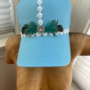 May include: Light blue baseball cap with white floral lace trim and a decorative center piece. The cap features a cluster of teal feathers and a silver-toned button. A stylish accessory for casual wear.
