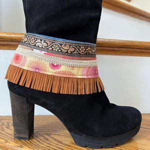 May include: Black suede boot with a decorative cuff. The cuff features a denim band, a patterned band with floral designs, a pink and yellow patterned band, a white lace trim, and a brown fringe. The boot has a wooden block heel.