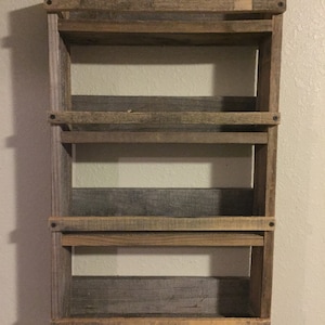 May include: A rustic wooden shelf unit with four shelves. The shelves are made of reclaimed wood and have a natural, weathered finish. The shelf unit is mounted to a wall and has a simple, farmhouse style.