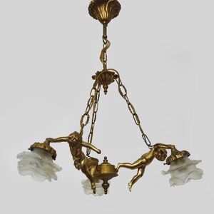 Puede incluir: L&aacute;mpara de ara&ntilde;a dorada ornamentada con tres pantallas de cristal esmerilado. La l&aacute;mpara est&aacute; adornada con figuras de querubines y eslabones de cadena decorativos, creando una pieza de iluminaci&oacute;n elegante y art&iacute;stica.