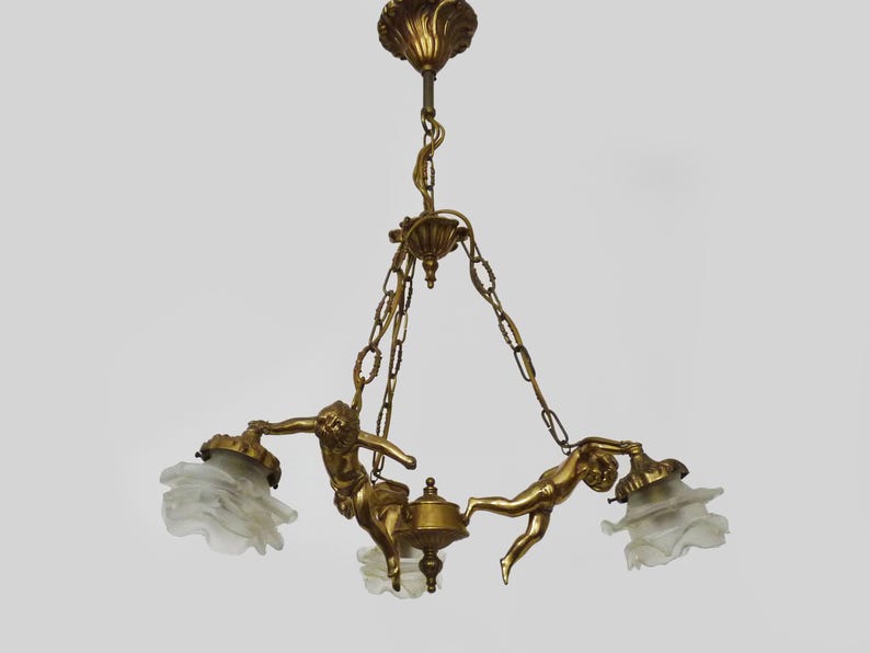Puede incluir: L&aacute;mpara de ara&ntilde;a dorada ornamentada con tres pantallas de cristal esmerilado. La l&aacute;mpara est&aacute; adornada con figuras de querubines y eslabones de cadena decorativos, creando una pieza de iluminaci&oacute;n elegante y art&iacute;stica.