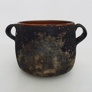 Op de afbeelding: Een ronde pot met twee handvatten en een donkere, verweerde zwarte buitenkant met zichtbare vlekken van bruine en grijze textuur. De binnenkant van de pot is glad en heeft een effen terracotta oranje kleur die contrasteert met de verouderde afwerking.