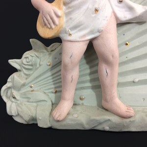 Puede incluir: Una figurita de porcelana de un querub&iacute;n tocando un instrumento musical, de pie sobre una base en forma de abanico verde y blanco con detalles de perlas.